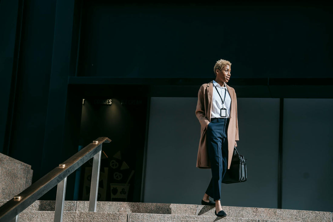 From below full body positive young female entrepreneur in stylish brown coat with hand in pocket and bag walking down stairs near office building