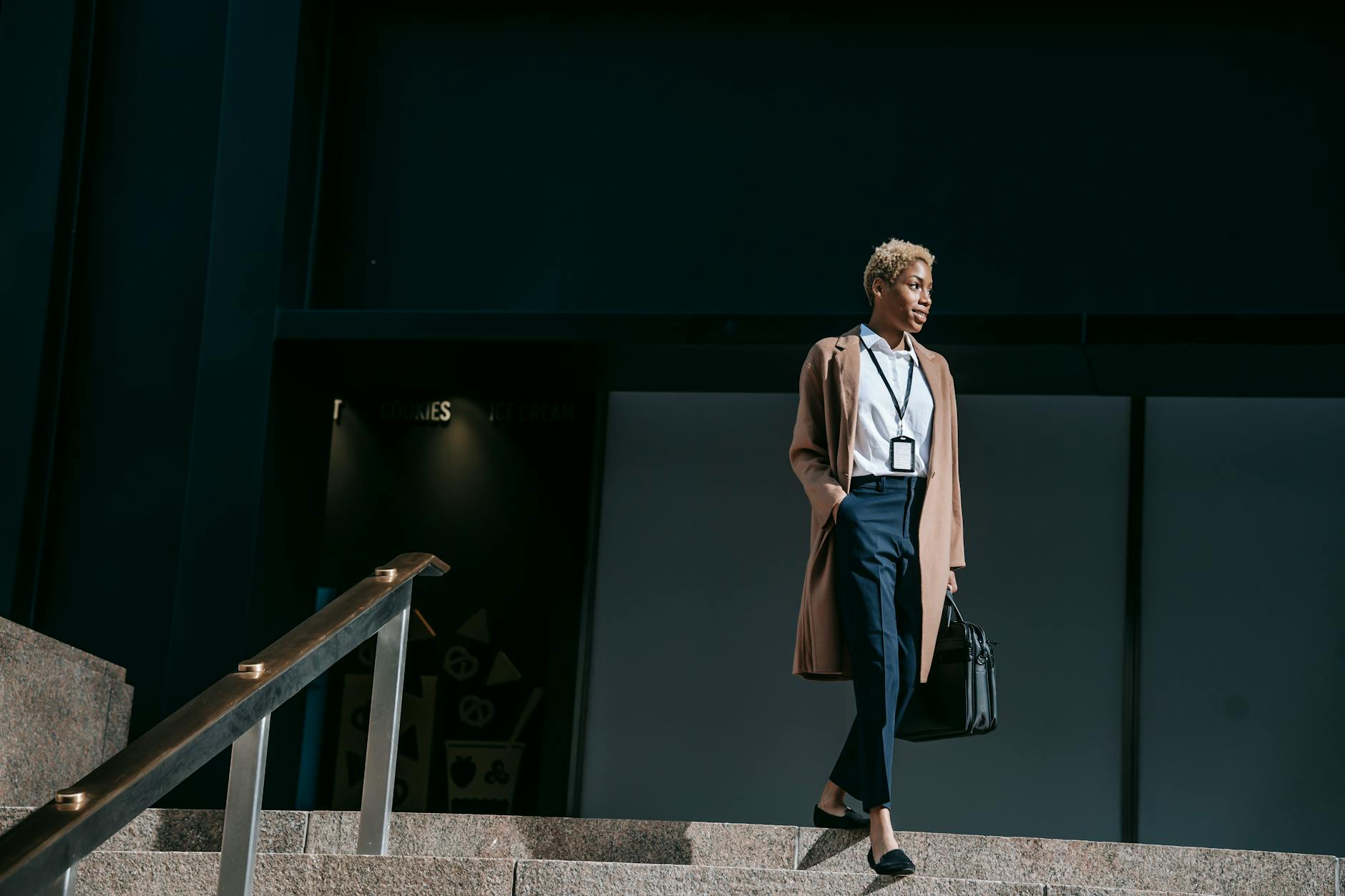 From below full body positive young female entrepreneur in stylish brown coat with hand in pocket and bag walking down stairs near office building